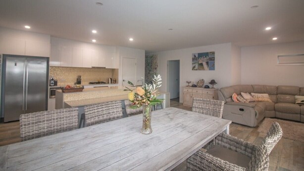 A modern kitchen with white cabinets, a stainless steel refrigerator, and a dining area with a wooden table and wicker chairs. - Azure on Bondi Place (Tweed)