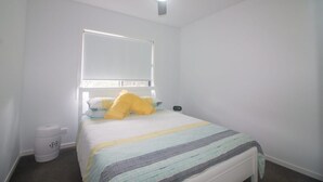 A bedroom with a bed, a ceiling fan, a window, and a trash can. - Azure on Bondi Place (Tweed)
