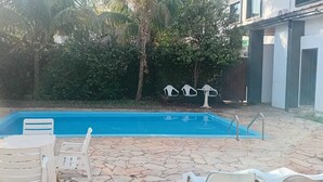 Una piscina rectangular con un patio de piedra, rodeada de sillas y mesas de plástico blanco.