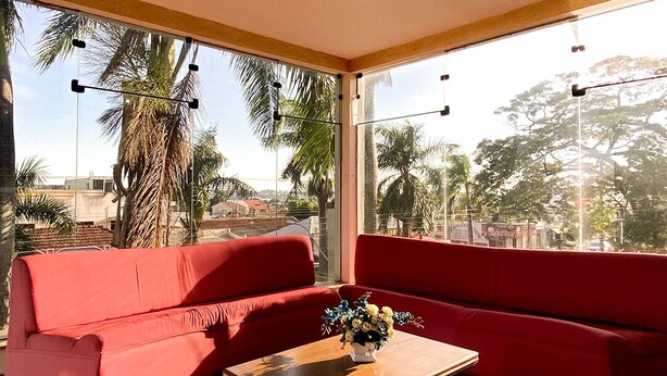 Habitación de hotel moderna con un sofá rojo, una mesa de centro de madera y ventanas amplias que ofrecen vistas a palmeras y edificios.