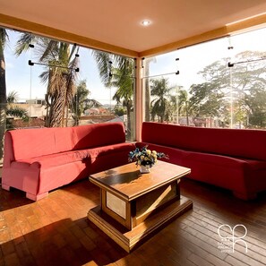 A modern hotel room with a red sofa set, a wooden coffee table, and large windows offering a view of palm trees and buildings.