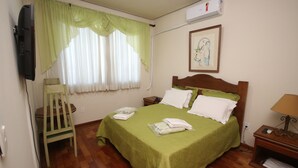 Una cama bien hecha con edredón verde, una mesita de noche de madera con una lámpara, una silla y un televisor montado en la pared.