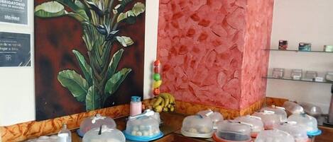 A reception desk with a large plant, a red textured wall, and several covered food containers.