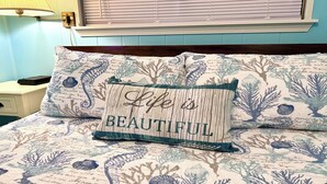 Uma cama com colcha estampada com flores, um travesseiro com a frase 'Life is Beautiful' e uma mesa de cabeceira com abajur e telefone.