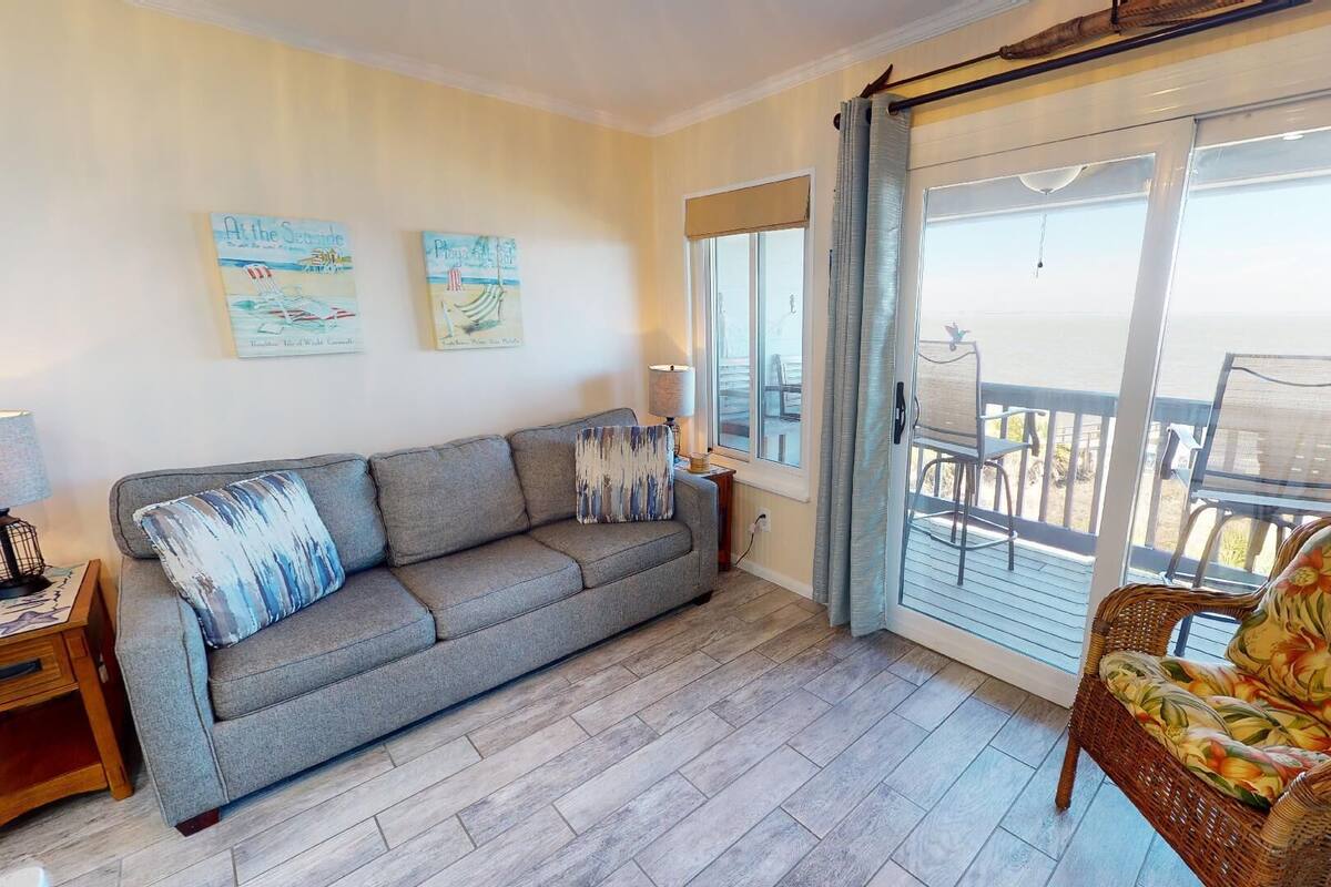 A living room with a grey sofa, a wicker chair, and a balcony with a view.