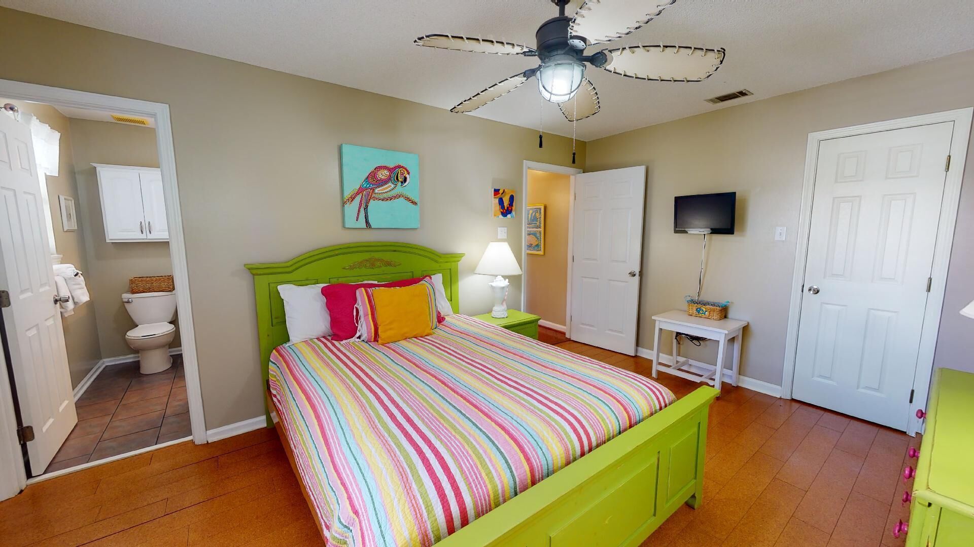 A bedroom with a green bed, a ceiling fan, a television, and a bathroom with a toilet and cabinet.