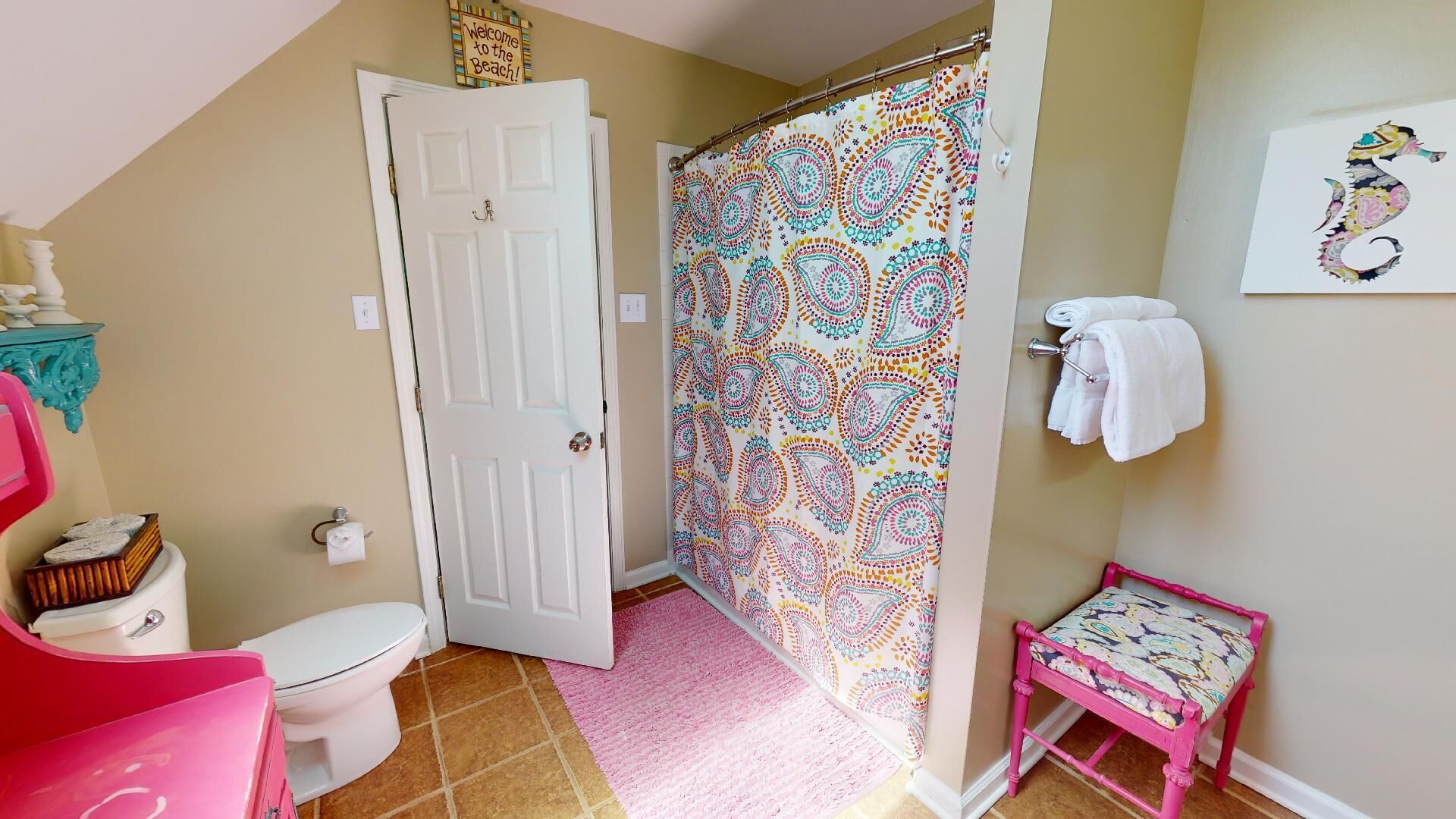 A bathroom with a white door, a colorful shower curtain, a pink chair, and a small bench.