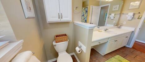 Une salle de bain avec des toilettes, un lavabo et un miroir. On y trouve un panier sur les toilettes, un porte-serviettes et un tapis vert sur le sol.