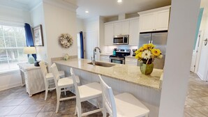 A kitchen with white cabinets, a stainless steel refrigerator, a microwave, and a sink. There are two white chairs and a small table with a lamp. A wreath hangs on the wall, and a vase with flowers is on the counter. - Serendipity by Tybee Vacation Rentals (Tybee Island)