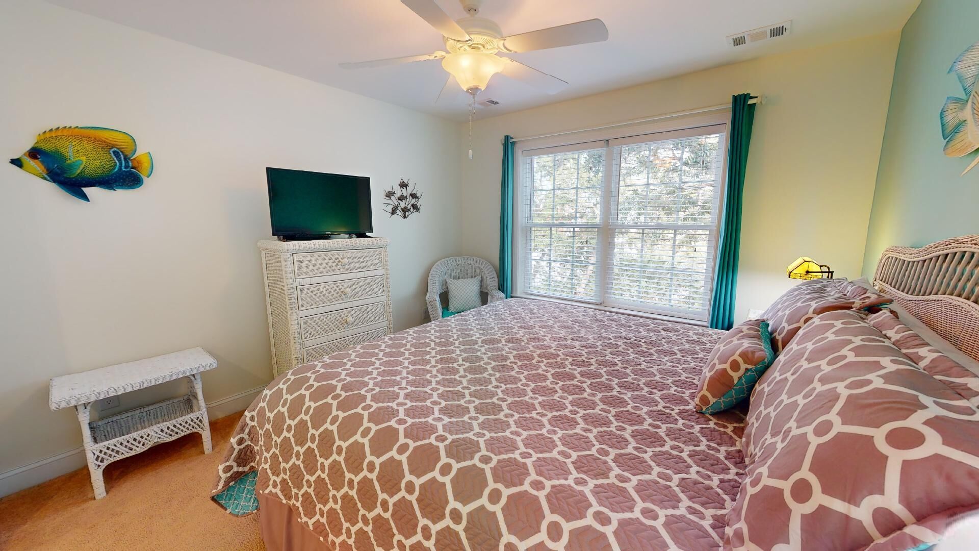 A bedroom with a bed, a ceiling fan, a television, a wicker chair, and a window with curtains.