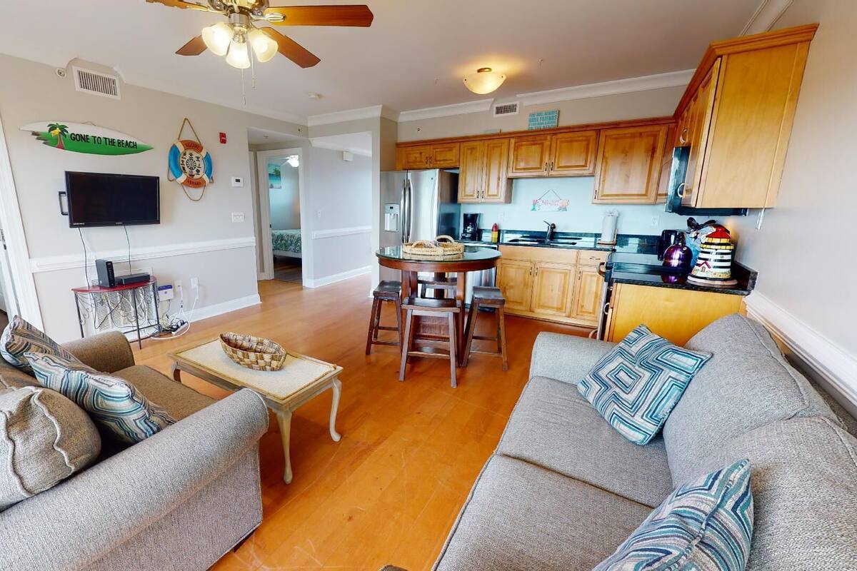 A spacious living room with a sofa set, a dining table with chairs, a kitchen with wooden cabinets, and a ceiling fan.