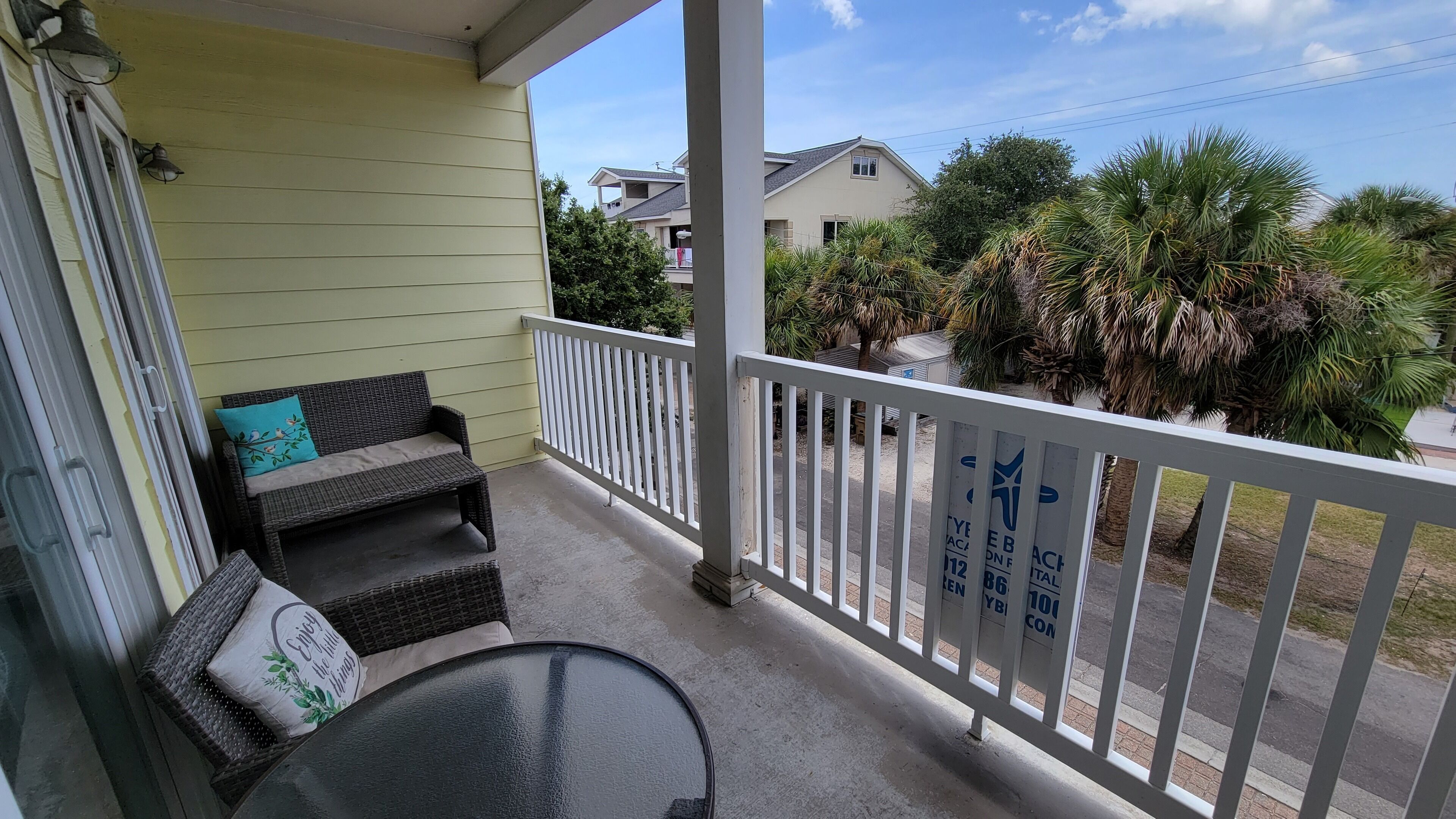 A balcony with a wicker sofa and a small table, overlooking a residential area with palm trees and houses.