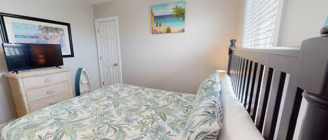 A bedroom with a bed, a television on a dresser, a door, and a painting of a beach scene.