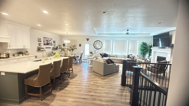 A spacious kitchen with a large island, a dining area with wicker chairs, a living room with a sofa and a fireplace, and a staircase with a railing.