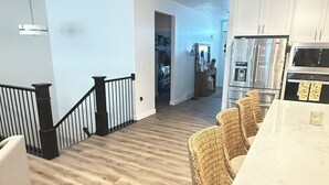 A spacious living area with a wooden floor, a dining area with wicker chairs, and a kitchen with white cabinets and stainless steel appliances.