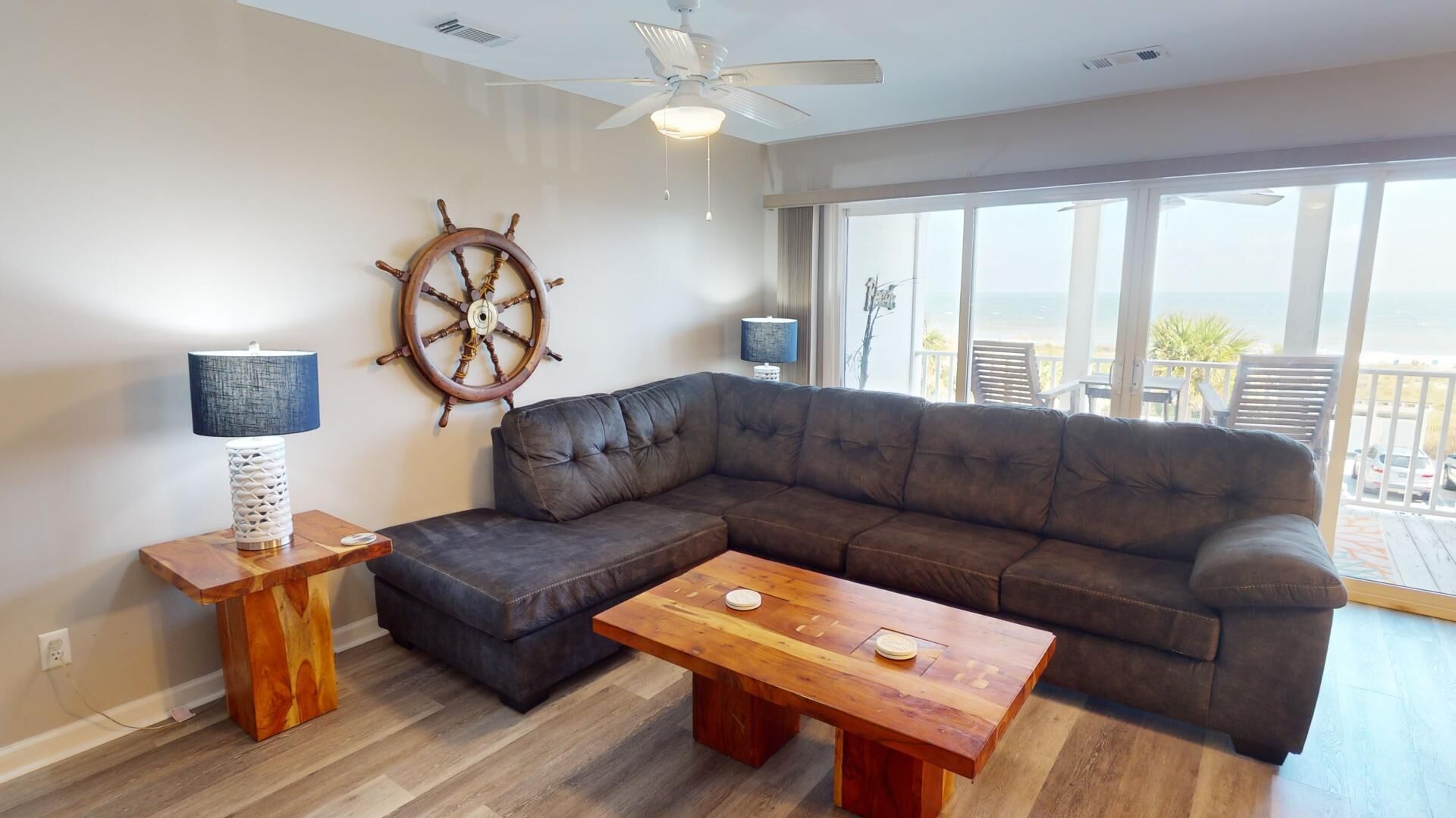 Una sala de estar con un sofá seccional marrón oscuro, una mesa de centro de madera, una decoración de timón de barco y un ventilador de techo.