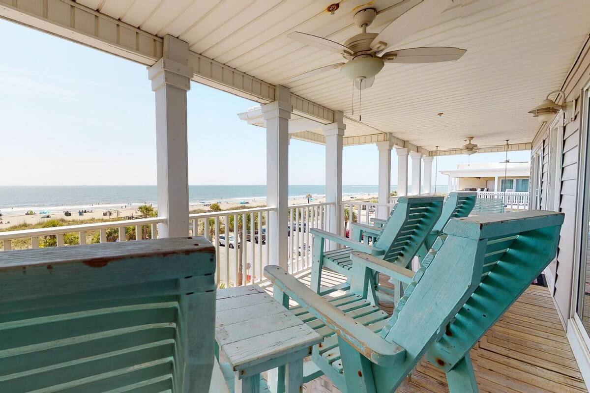 A covered porch with a view of the beach, featuring wooden chairs and a ceiling fan.