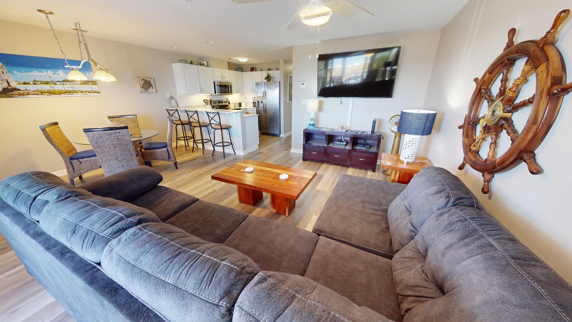 A living room with a large gray sofa, a wooden coffee table, and a TV mounted on the wall. There is a kitchen in the background with white cabinets and a dining area with chairs.