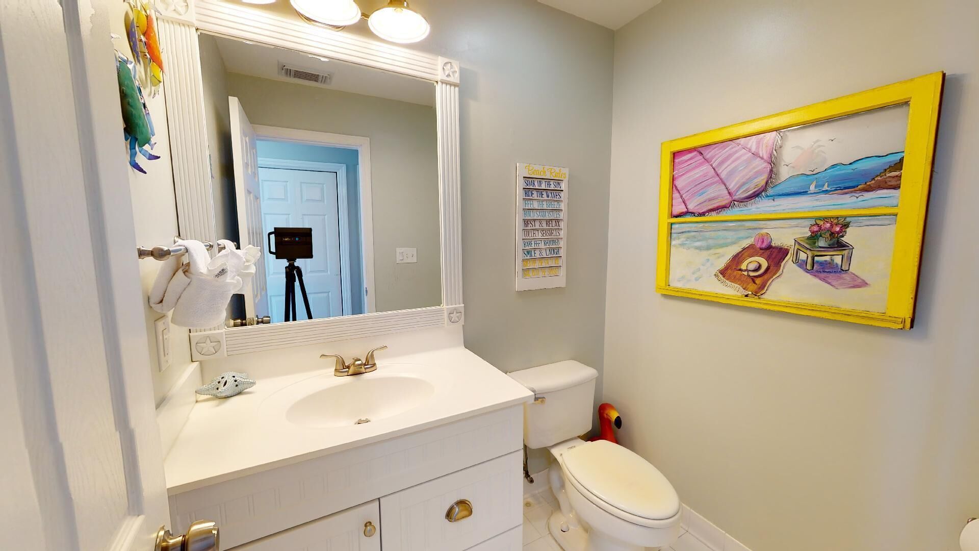 A bathroom with a white sink, a toilet, and a large mirror. There is a framed painting on the wall and a sign with a list of items.