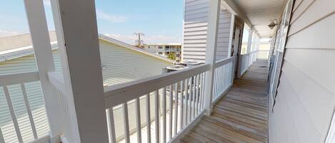 A wooden deck with white railings, leading to a covered porch area with a view of neighboring buildings.
