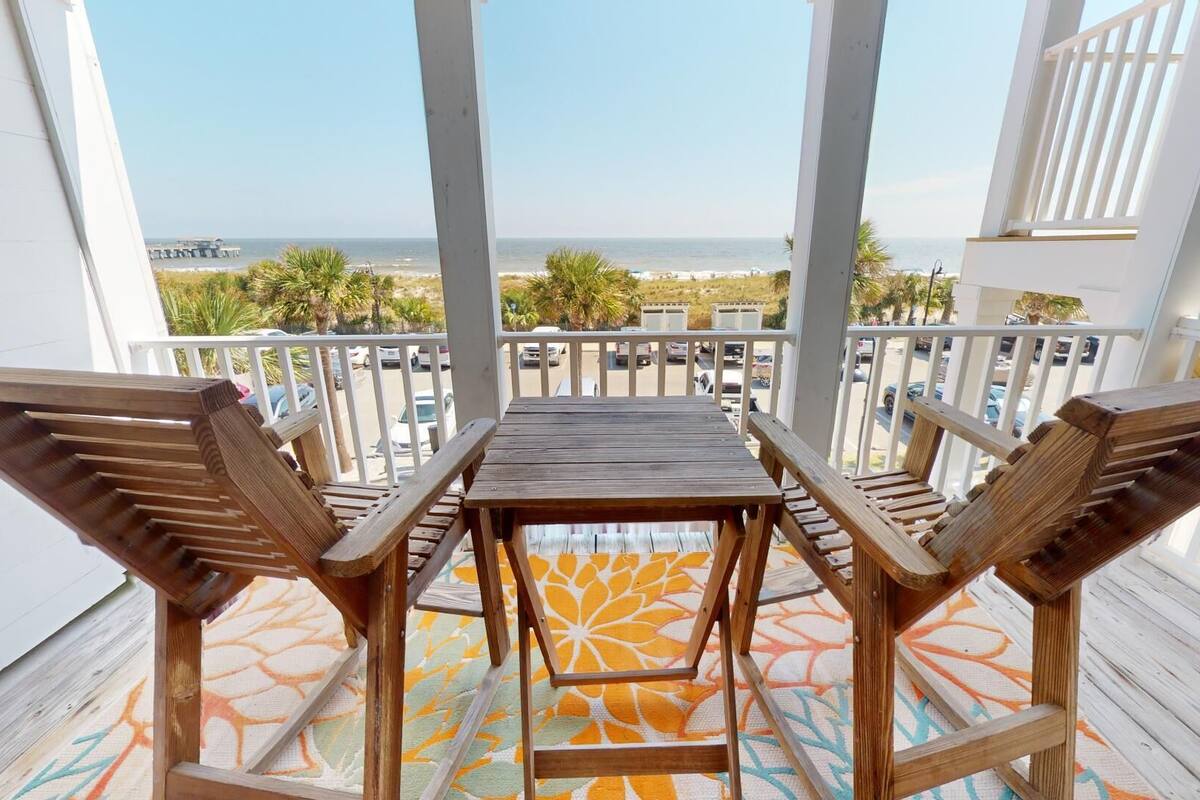 A wooden deck with a table and chairs, overlooking a beach with palm trees and a pier.