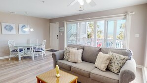 En stue med en grå sofa, et glasbord med hvide stole og et stort vindue med udsigt til en balkon og palmer.