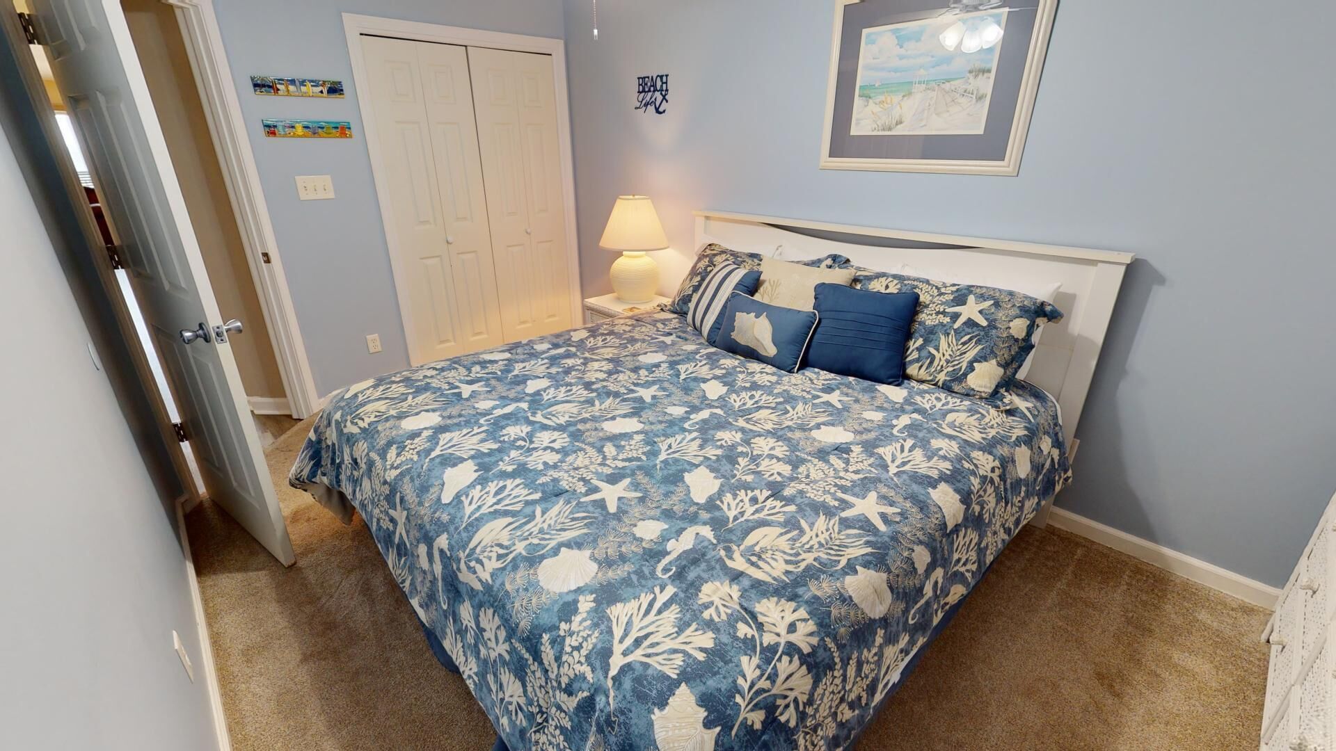 A bedroom with a bed, a nightstand, a lamp, a door, and a wall with a picture and a sign.