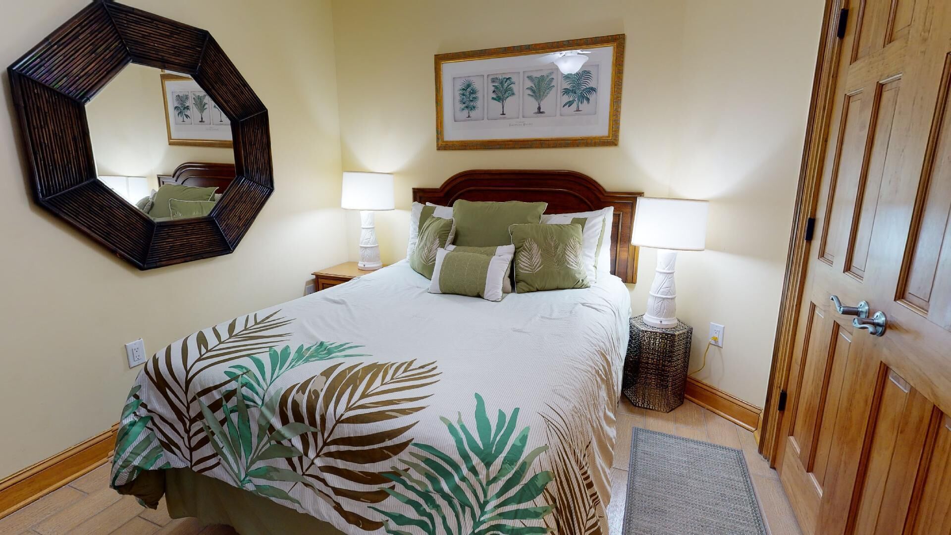 A hotel room with a large bed, two bedside lamps, a mirror, a framed botanical print on the wall, and a wooden door.