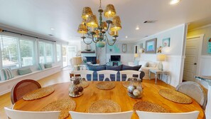 A spacious living room with a large wooden dining table surrounded by woven placemats and white chairs. A modern chandelier hangs from the ceiling, and the room features a cozy seating area with a blue and white patterned sofa. - The Island Star by Tybee Vacation Rentals (Tybee Island)