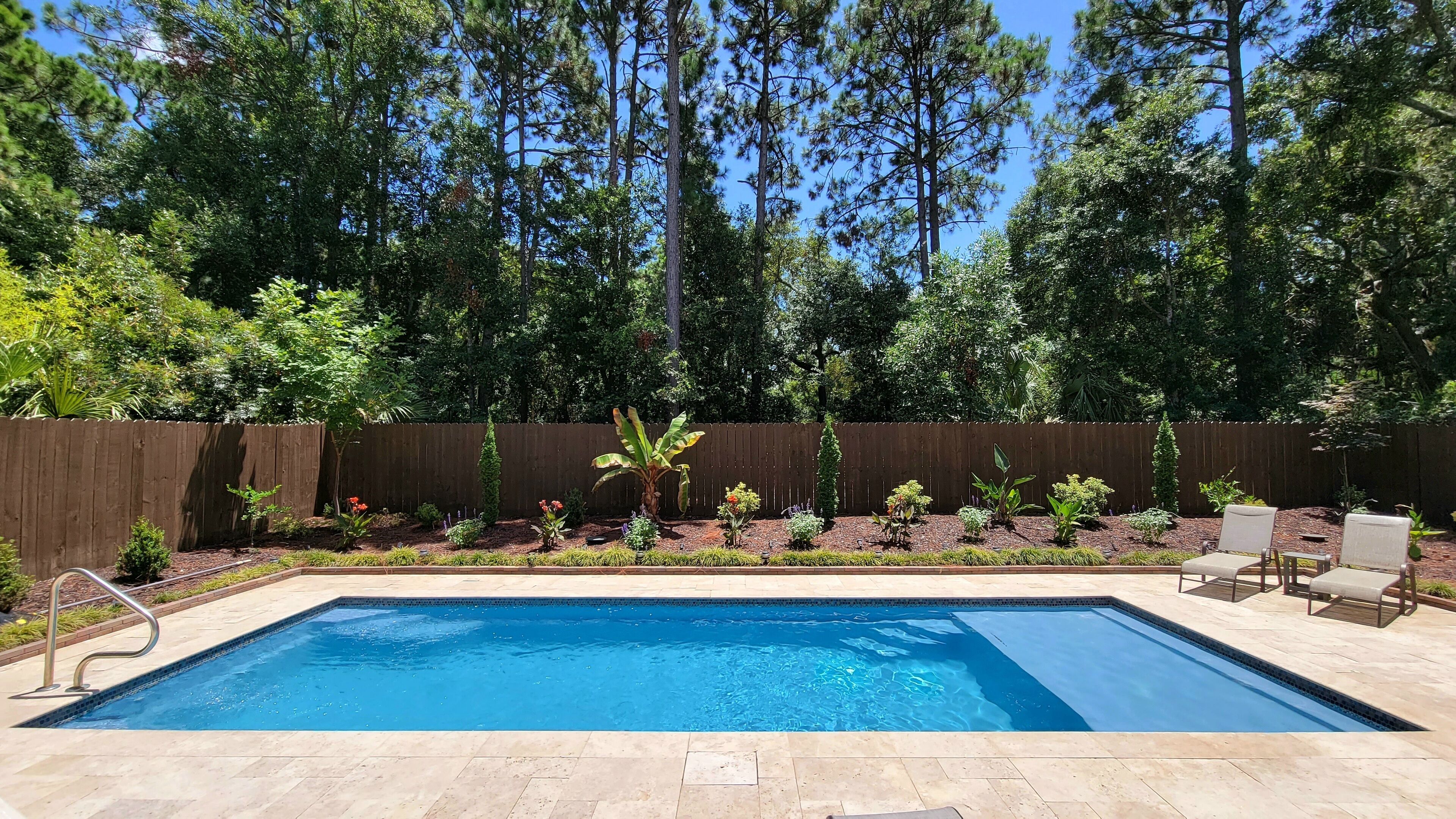 A rectangular swimming pool with a wooden deck surrounding it, a wooden fence, and various plants and shrubs.