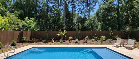 A rectangular swimming pool with a wooden deck surrounding it, a wooden fence, and various plants and shrubs.