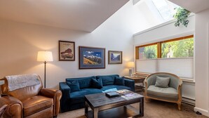 Uma sala de estar com uma poltrona de couro marrom, um sofá azul, uma mesa de centro de madeira e uma poltrona cinza claro. Há quadros nas paredes e uma janela com persianas.