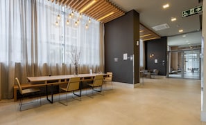 Un moderno vestíbulo de hotel con un mostrador de recepción de madera, sillas y un gran ventanal con cortinas traslúcidas.