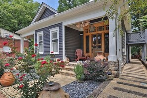 Une maison avec le numéro « 506 » au-dessus de la porte, un porche avec une chaise et un jardin où poussent différentes plantes, ainsi qu’un abreuvoir pour les oiseaux.
