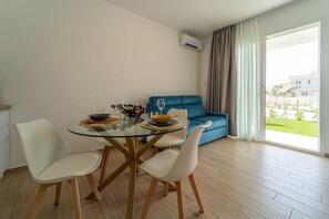 A dining area with a glass table, white chairs, a blue sofa, and a view of a building through a large window. - GuestHost - Charming 40 sqm apartment, able to accommodate up to 4 people, located within the renowned Borgo 84 residence, featuring two swimming pools and a bike path. The property is located just 3 km from the center of Marzamemi, one of the most fasci (Pachino)