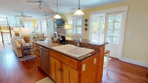 A spacious kitchen with wooden cabinets, a granite countertop, and a double sink. There is a dining area with a table and chairs, and a living room with a sofa and a television.