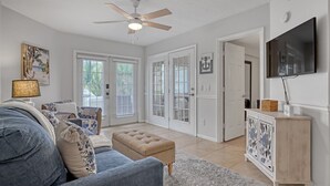 A spacious living room with a ceiling fan, a large TV mounted on the wall, a couch, a coffee table, and a cabinet. - Heated pool, hot tub, and private beach access! (Cape Canaveral)