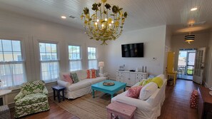 A spacious living room with white walls, a large chandelier, and a television mounted on the wall. - Twelve Palms Cottage by Tybee Vacation Rentals (Tybee Island)