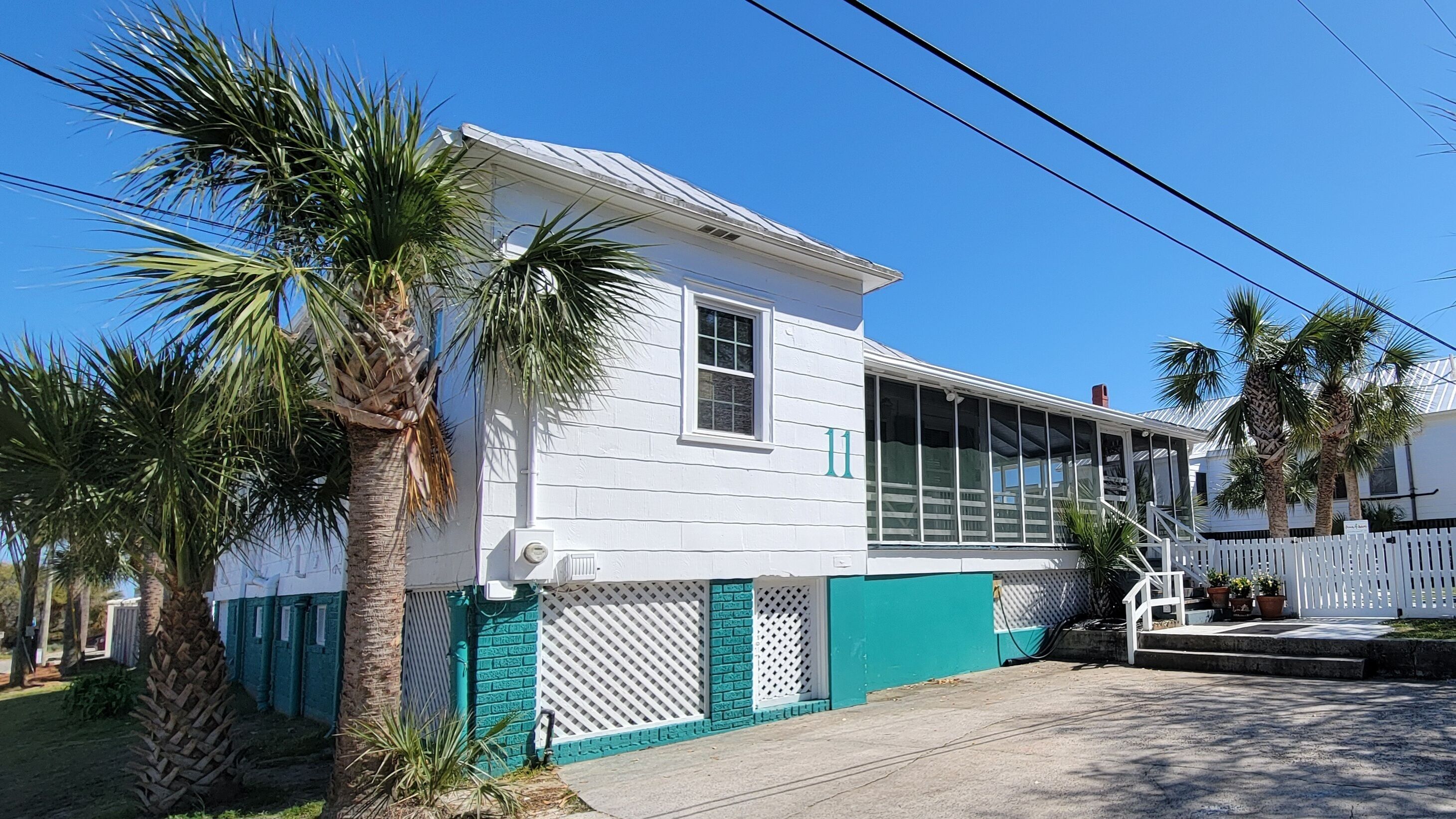 A white building with a large window and a door, surrounded by palm trees and a white picket fence.