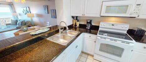 A kitchen with granite countertops, stainless steel sinks, white cabinets, a white stove, and a microwave.