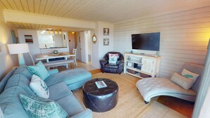 A living room with a sofa, ottoman, and armchair, a dining area with a table and chairs, and a TV on a wooden wall. - Tybee Lights 112C by Tybee Vacation Rentals (Tybee Island)