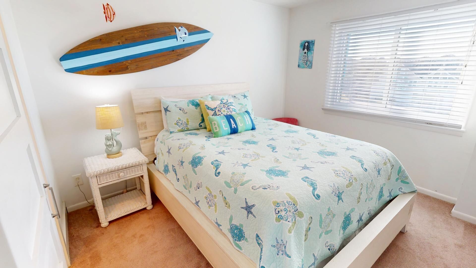 A bedroom with a bed, a nightstand, a surfboard decoration, and a window with blinds.