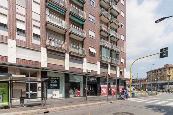 A multi-story building with balconies, a bus stop, and storefronts including a pizza place and a clothing store.