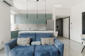 A modern living room with a blue sofa, teal cabinets, and a kitchen area with a refrigerator and sink.