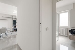 A modern, minimalist room with a white wardrobe, a dining area with a table and chairs, and a view of a bathroom with a toilet and radiator.