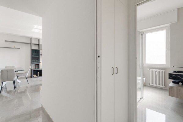 Una habitación moderna y minimalista con un armario blanco, una zona de comedor con mesa y sillas, y vistas a un baño con inodoro y radiador.