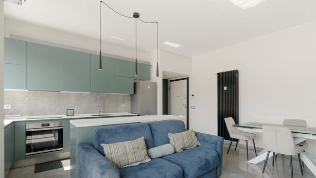 Una cocina moderna con armarios color esmeralda, un sofá azul con almohadones a rayas, y un comedor con mesa y sillas blancas.