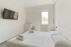 A modern hotel room with a large bed, a flat-screen TV mounted on the wall, a window with blinds, and a radiator.