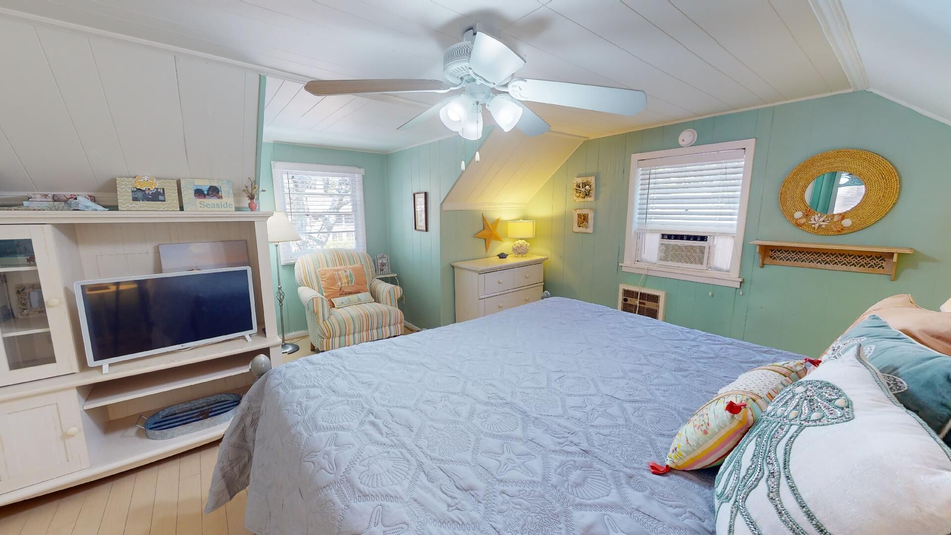 Une chambre à coucher comprenant un lit, un téléviseur, un ventilateur de plafond, une commode, une chaise et une fenêtre avec des stores.