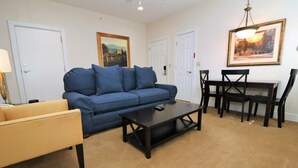 A living room with a blue sofa, a wooden coffee table, a yellow armchair, a dining table with chairs, and two framed pictures on the walls.